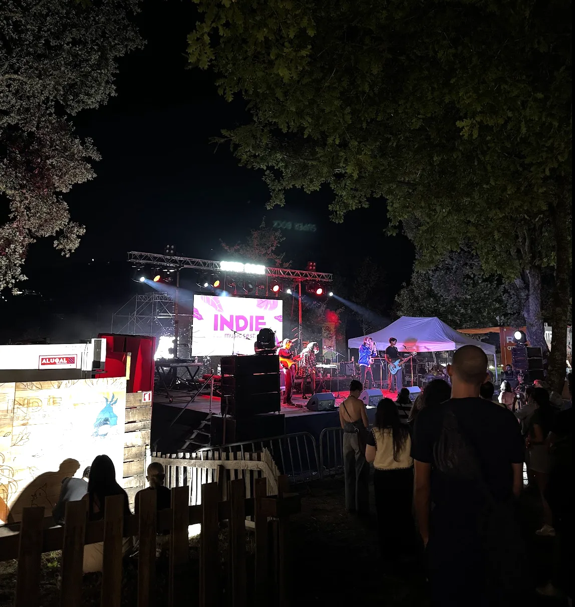 Banda em palco e público a assistir a um concerto entre as árvores do Indie Music Fest à noite