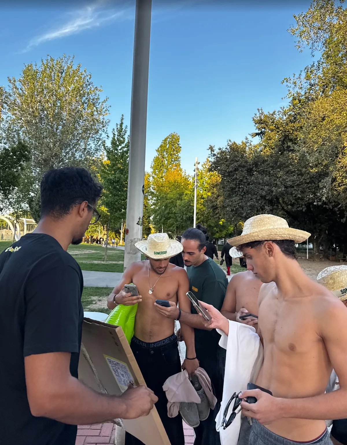 Estudiantes con sombrero descargando la app en el móvil junto al cartel con el código QR, camino de las piscinas