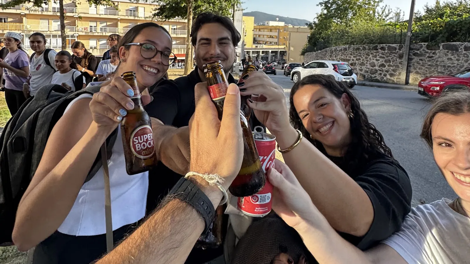 Cuatro personas brindando con botellas de cerveza y una lata al final de la tarde, junto al campus de la UMinho