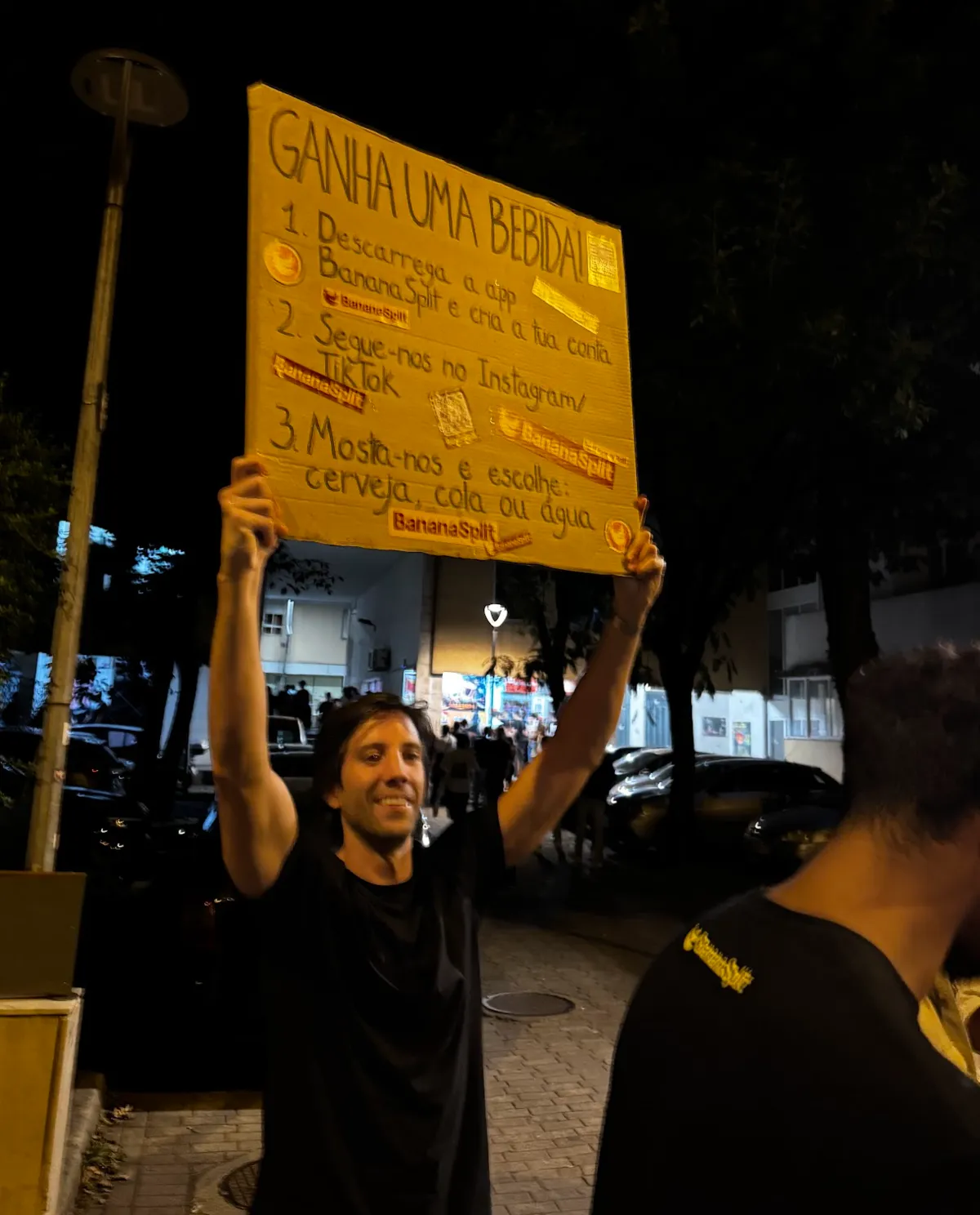 Nuno levantando el cartel «¡Gana una bebida!» por encima de la cabeza, en la calle de noche