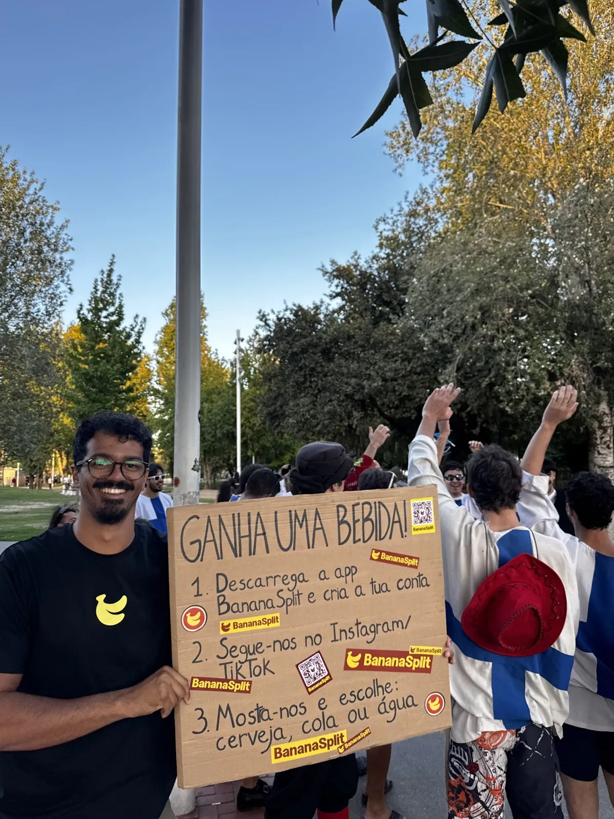 Leo sosteniendo el cartel «¡Gana una bebida!» en el parque de la Rodovia, con un grupo de estudiantes celebrando detrás